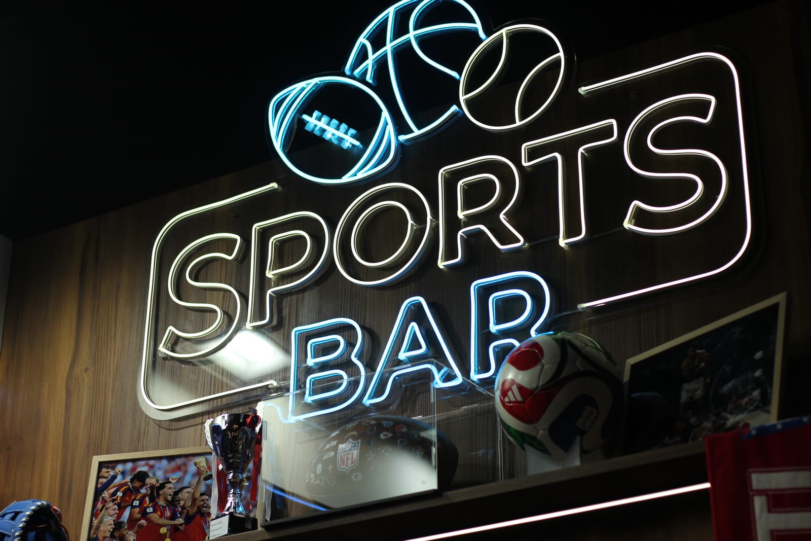 The neon Sports Bar sign above the bar on the 2Coders stand at IBC 2026, with a trophy, an NFL helmet and a match ball on the shelf below it.
