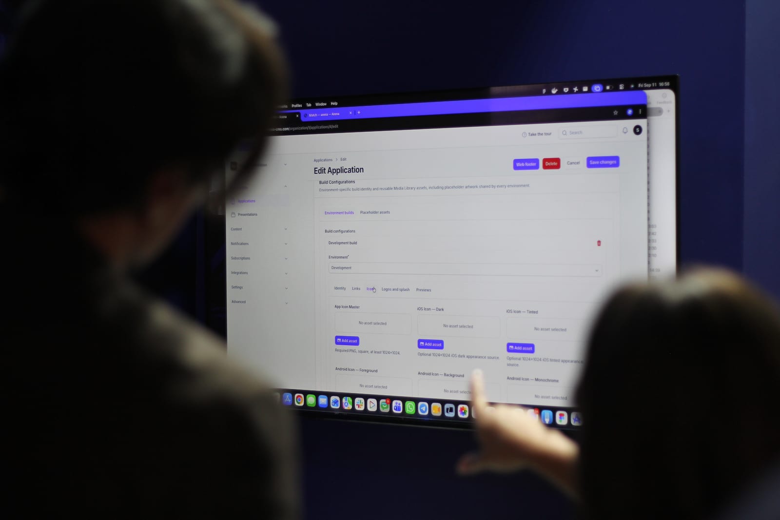 Two people at the stand looking at the Arena CMS on a laptop, on the Edit Application screen showing build configurations and app icon assets.